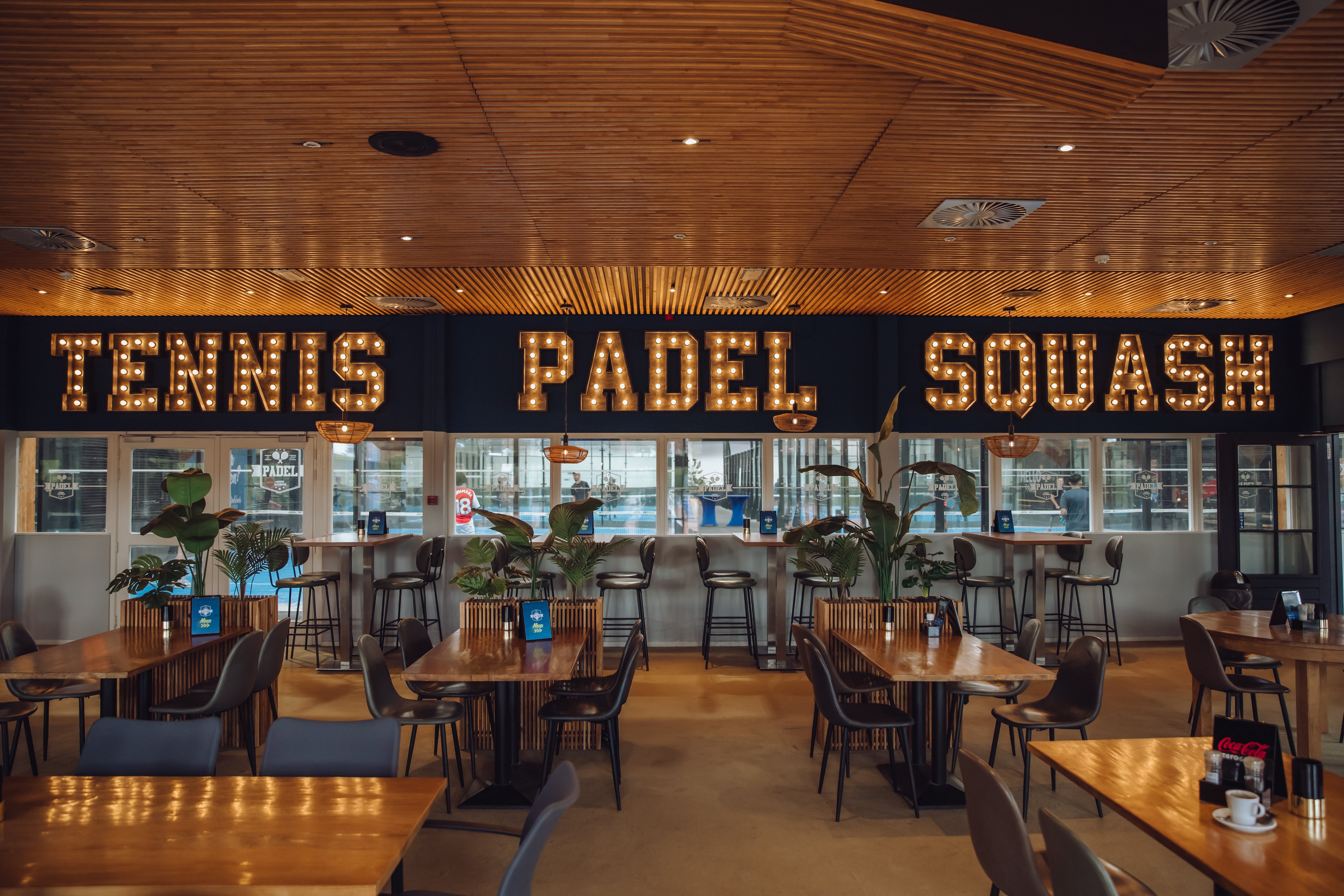 Sfeervol interieur van de sportbar bij Dekker Zoetermeer, met houten tafels, comfortabele stoelen en verlichte letters Tennis, Padel en Squash. Vanuit de sportbar hebben gasten direct uitzicht op de indoor padelbanen.