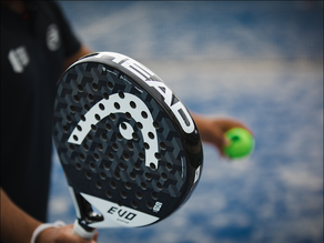 Close-up van een padelracket met een groene padelbal in de hand van een speler op de blauwe padelbaan bij Dekker Zoetermeer.
