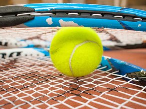 Close-up van een felgele tennisbal tussen twee blauw-zwarte tennisrackets op een roodkleurige tennisbaan bij Dekker Zoetermeer.