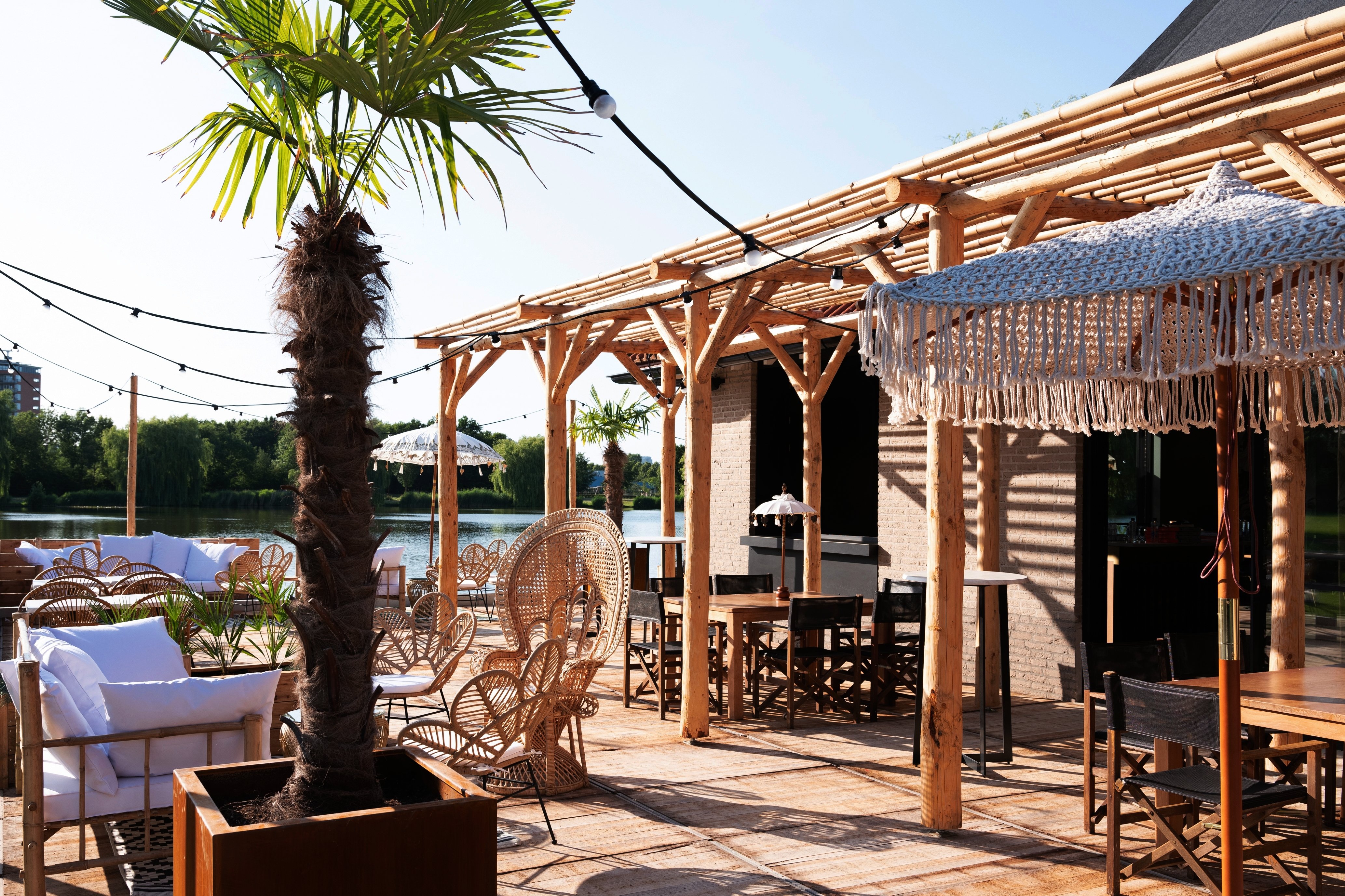 Sfeervol terras aan het water bij Dekker Zoetermeer met palmbomen, houten pergola, rotan loungemeubels en gezellige zitplekken in de zon.