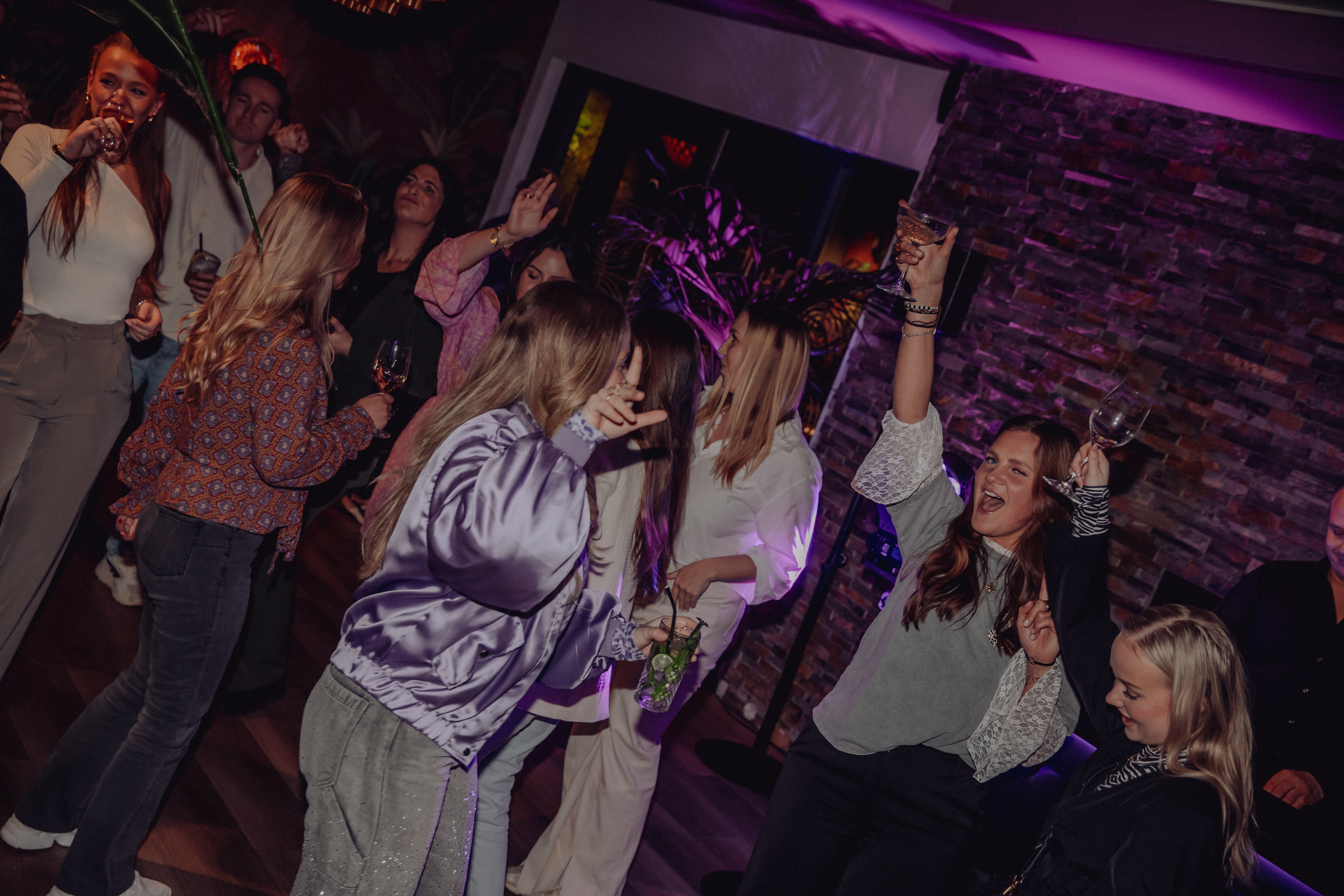 Dansende gasten met drankjes en opgeheven armen tijdens een feest met paarse verlichting